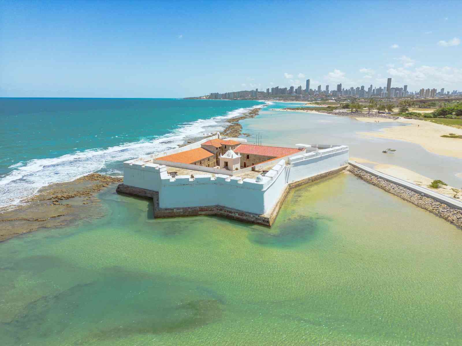Vista aérea do Forte dos Reis Magos em Natal, Rio Grande do Norte, Brasil. A fortaleza histórica em formato de estrela, com paredes brancas e telhados de terracota, está estrategicamente localizada na foz do Rio Potengi, cercada por águas cristalinas do oceano Atlântico e uma praia de areia clara. Ao fundo, a silhueta da cidade de Natal se estende sob um céu azul claro e ensolarado.