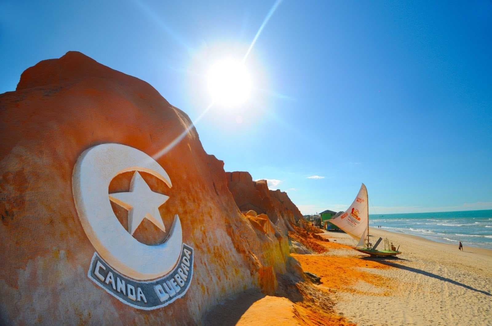 Símbolo icônico da lua e estrela de Canoa Quebrada esculpido nas falésias de areia avermelhada, com uma jangada tradicional de vela branca na praia de areia clara, sob um céu azul intenso e um sol brilhante. Ao fundo, o mar azul-turquesa e a linha do horizonte.