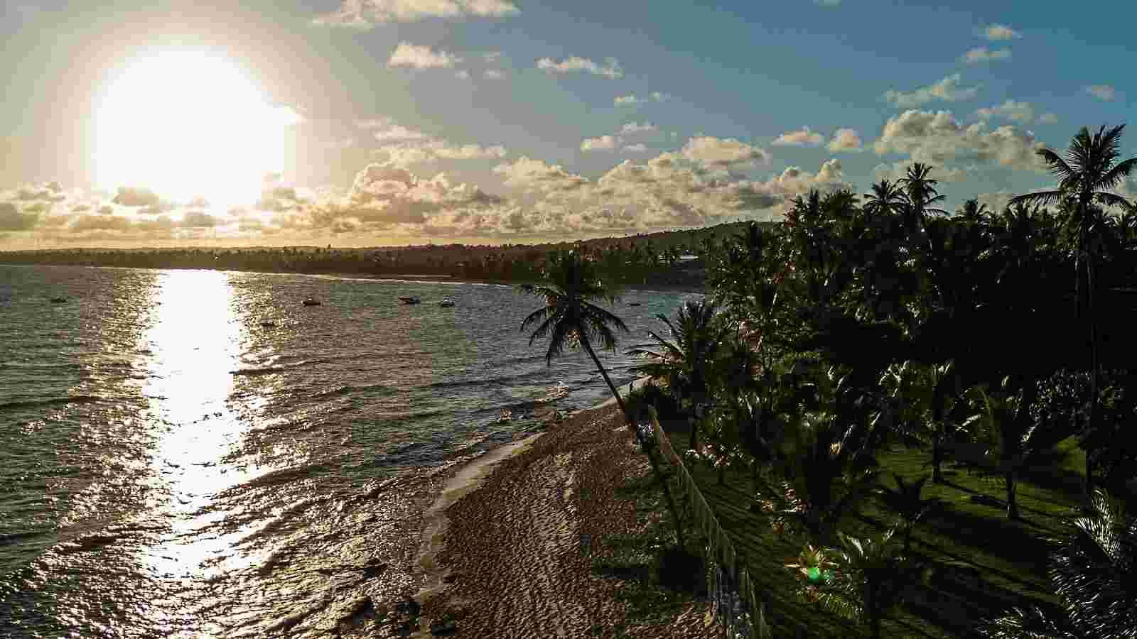 Vista aérea de um pôr do sol vibrante sobre uma praia tropical com areia dourada e coqueiros exuberantes. O sol intenso reflete-se no mar calmo, criando um caminho dourado na água, enquanto pequenos barcos flutuam ao longe. O céu está parcialmente nublado com tons de azul e laranja, e a vegetação densa da costa completa a paisagem paradisíaca.