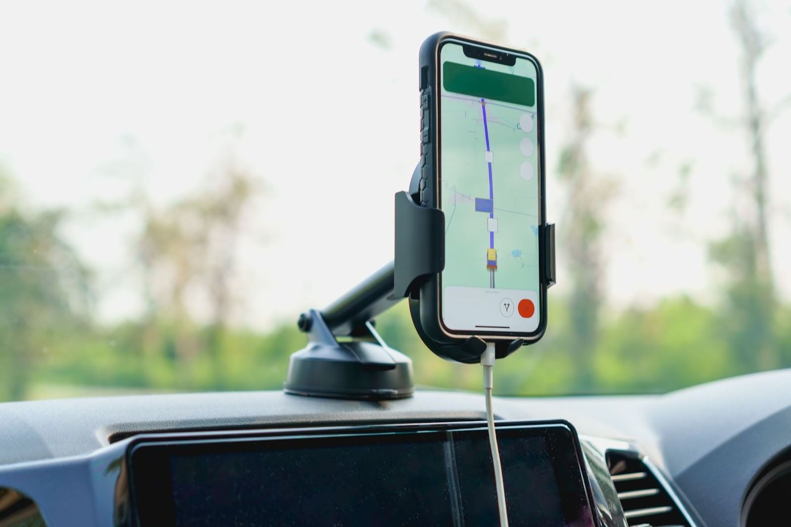 Smartphone preto em um suporte veicular no painel de um carro, exibindo um aplicativo de navegação GPS com uma rota em andamento. O celular está conectado a um cabo branco de carregamento, com o fundo desfocado de árvores e céu, indicando uma viagem ou percurso.